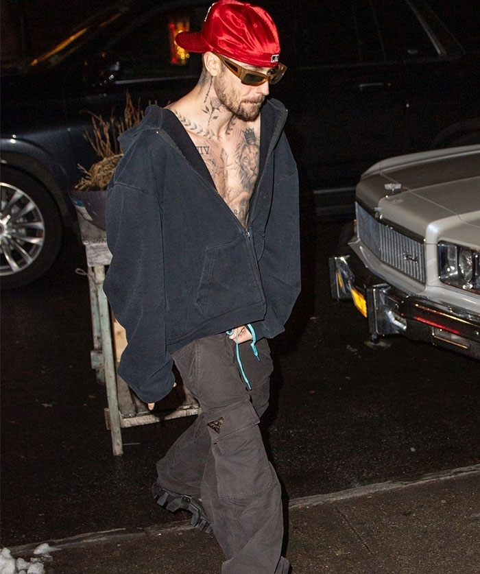 Man in urban attire with tattoos walks by a car at night, wearing a red cap and sunglasses, capturing a casual moment.