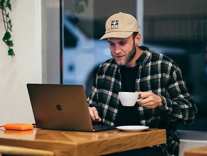 Man in a plaid shirt and hat, seated with a laptop and coffee, symbolizing taking time for himself.