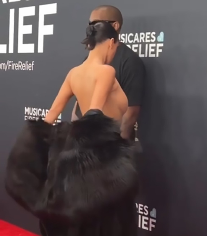 Bianca Censori on the red carpet, wearing a black outfit, with Kanye West in the background.