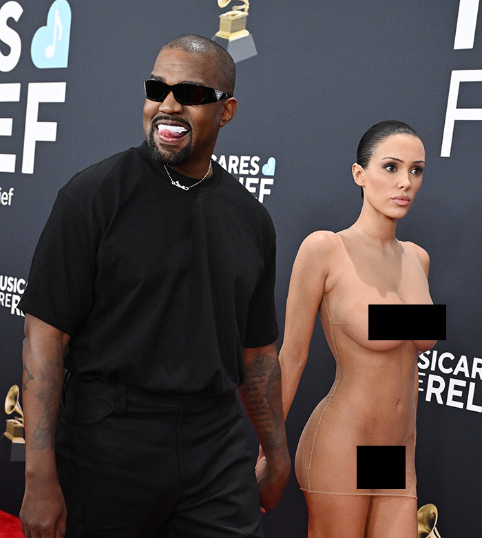 Kanye West in black attire and sunglasses attends Grammys with Bianca Censori in a sheer outfit, posing on the red carpet.