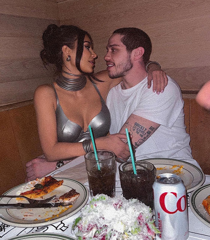 Kim Kardashian and Pete Davidson sitting closely at a restaurant table, one with visible tattoos, pizza and drinks on the table.