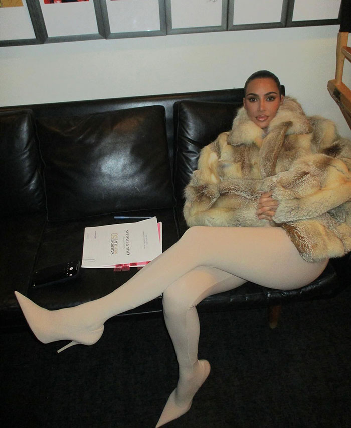 Woman in fur coat seated on a black couch next to documents, embodying modern style and elegance indoors.