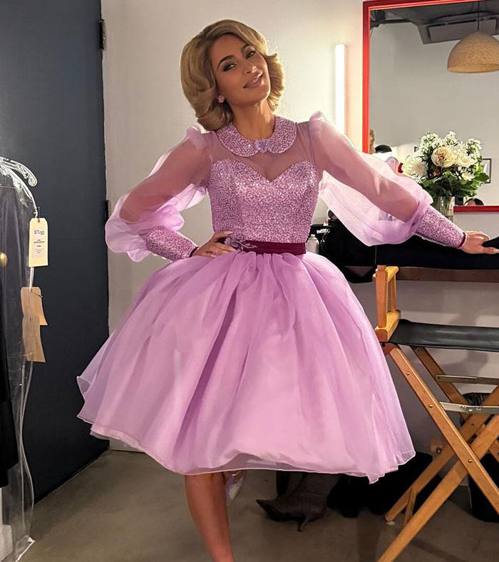 Woman in a lilac dress posing in a dressing room setting.