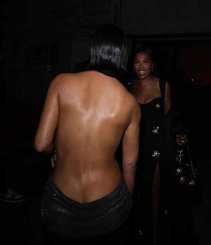 Two women in elegant evening attire at a social event, one in a backless dress, smiling.