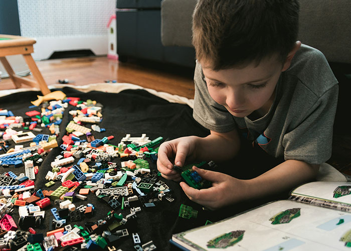 Child playing with toy bricks on the floor, absorbed in building from a book.