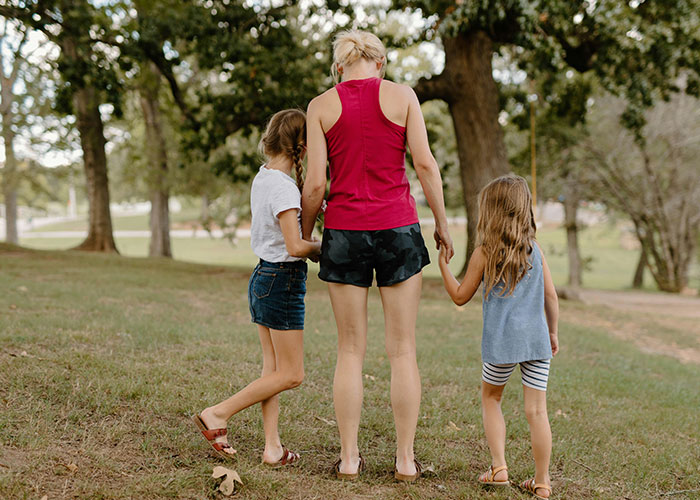 A mother with two kids walking in a park, holding hands, surrounded by trees.