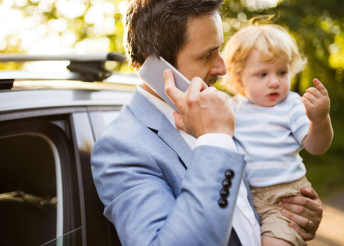 Man in blue suit holding a child, talking on phone outdoors, illustrating family dynamics.