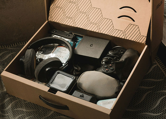 Open box with electronics and gaming items, including a headset and controllers, on a textured surface.