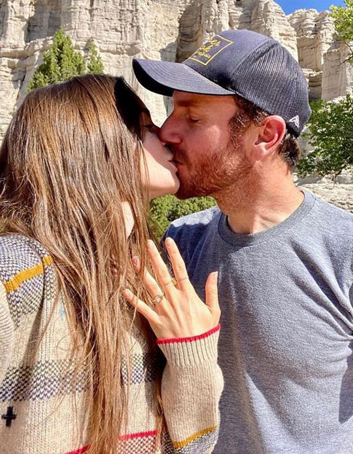 Couple kissing outdoors, woman showing engagement ring against rocky backdrop.