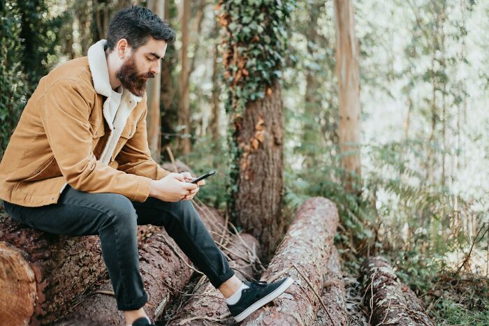 Man in a brown jacket sitting on logs in a forest, looking at his phone, offering a fresh perspective on the world.