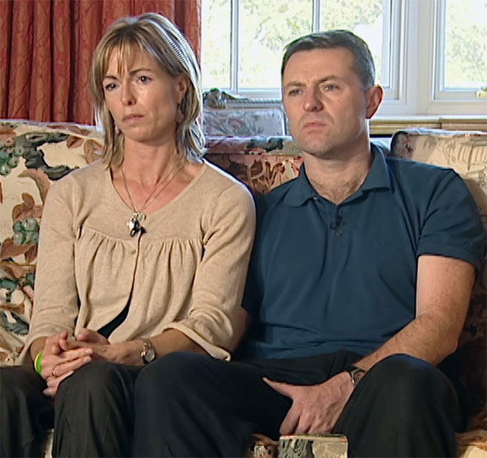 Man and woman sitting together on a sofa, looking serious, in a room with floral curtains and a window behind them.