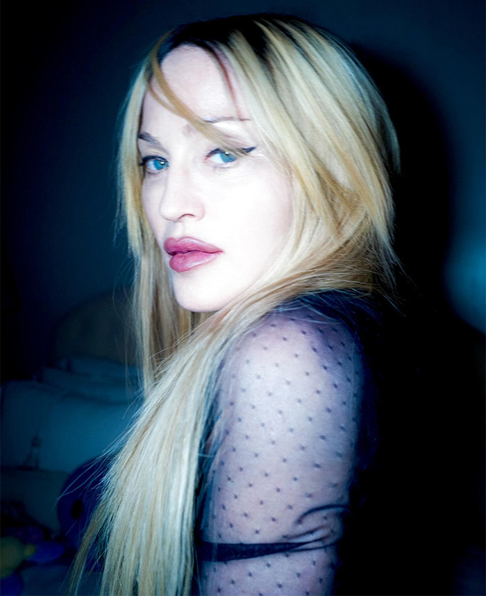 Blonde woman with long hair in a sheer polka dot top, looking over her shoulder under cool lighting.