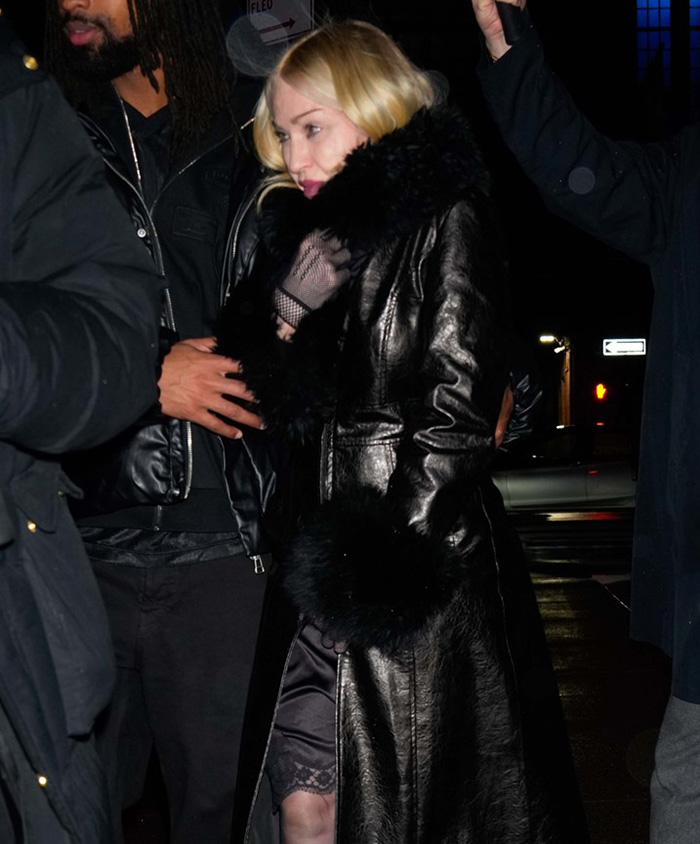 A woman in a black leather coat steps out in NYC at night, surrounded by others.