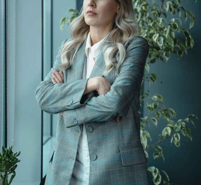 Woman in a plaid blazer stands with arms crossed, looking out of a window, contemplating something private on her laptop.