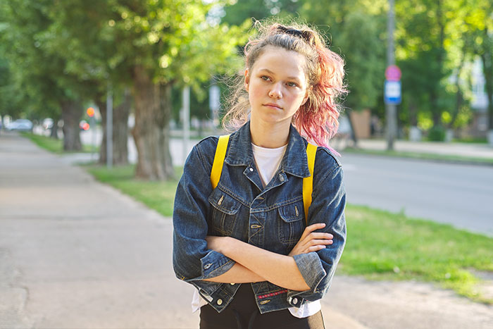 Teen girl with a denim jacket stands on a sidewalk, arms crossed, expressing defiance, related to pathological lies topic.