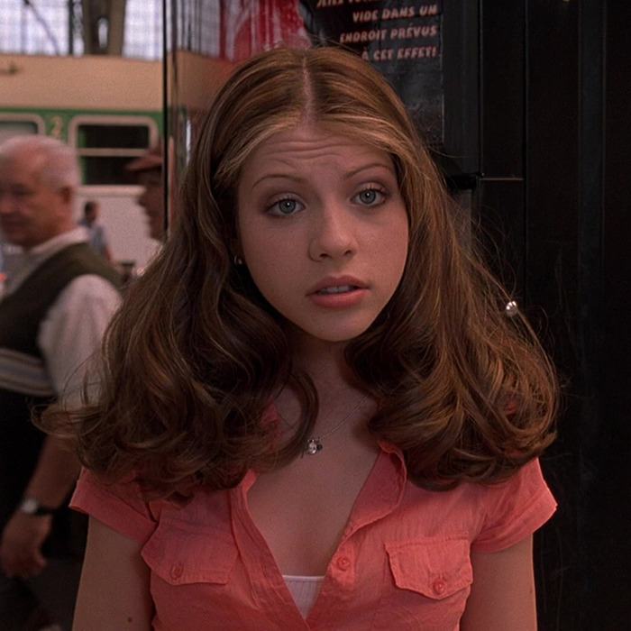 Michelle Trachtenberg with long hair wearing a pink top, standing indoors.