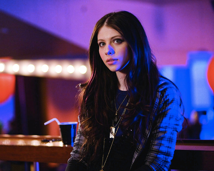 Michelle Trachtenberg sitting in a dimly lit setting with blue lights, wearing a layered outfit in a casual ambiance.