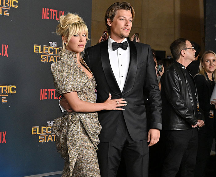 Millie Bobby Brown at The Electric State film premiere, wearing a glamorous gown with co-star in a suit on the red carpet.