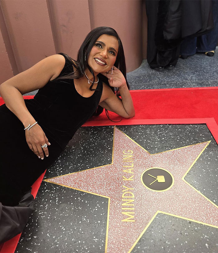 A woman in a black dress lies beside a Hollywood star on the Walk of Fame, smiling for the camera.