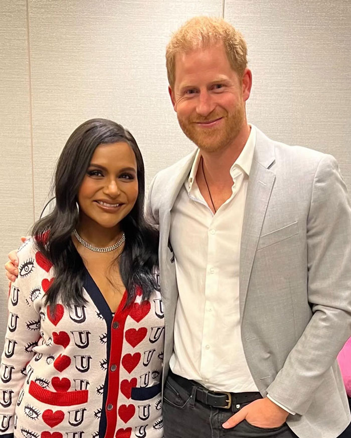 Two people smiling, one in a patterned sweater and the other in a light gray jacket, standing together indoors.