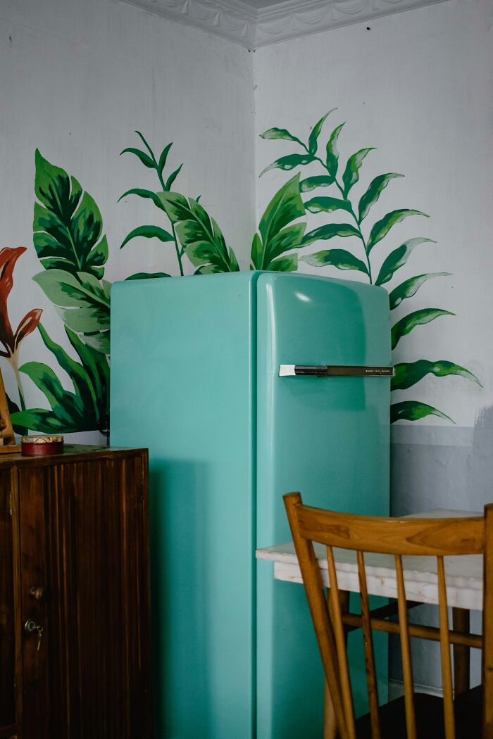 A vintage teal fridge in a rustic kitchen setting with leafy wall decor, highlighting boomer nostalgia.