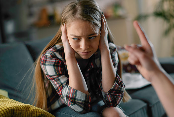 Teen girl looking frustrated, covering ears while being scolded, surrounded by home setting.