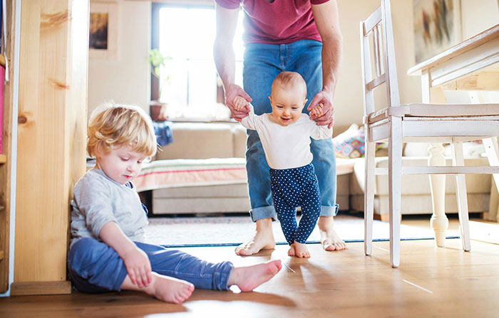 Parent helping baby walk while toddler sits nearby, showcasing sibling dynamics and parenting challenges.