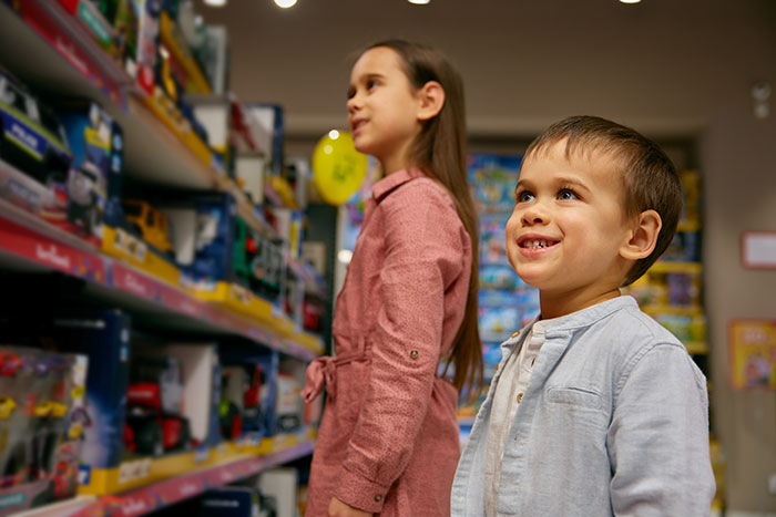 Kids in a store looking at toys on shelves, with a boy smiling and a girl observing in a pink dress.