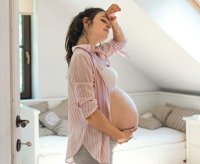 Pregnant woman stands thoughtfully in a cozy bedroom, touching her belly.