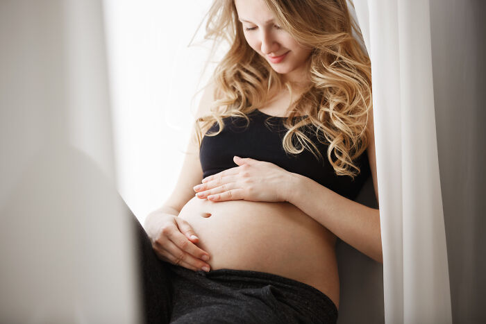 Pregnant woman in a peaceful moment by the window, touching her belly with a gentle smile.