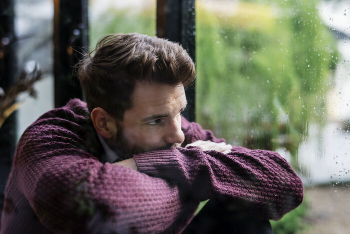 Man in maroon sweater looking thoughtful by a rainy window, balancing job and house move challenges.