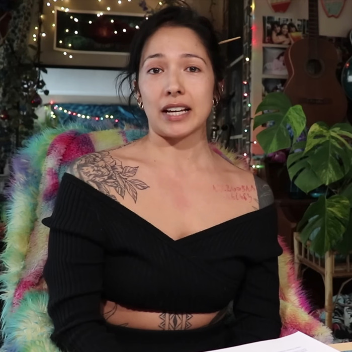 Woman discussing BookTube crisis, sitting in a cozy room with plants and lights, wearing a black top.