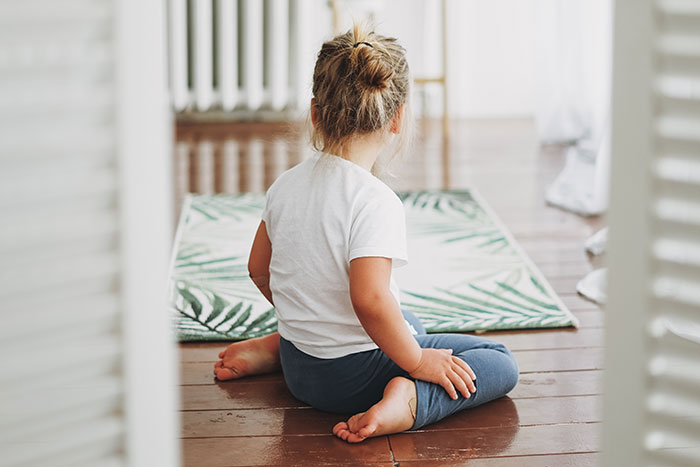 Child sitting on a yoga mat, facing away, in a calm room with natural light, suggesting a serene setting.