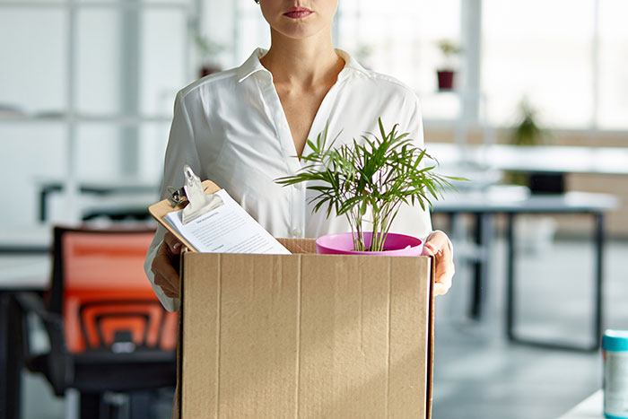 Woman in an office holding a box with a plant and clipboard, illustrating a workplace shift conflict.