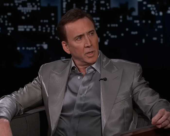 Nicolas Cage in a silver suit during a TV interview, seated and speaking animatedly.