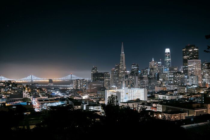 Skyline silhouette of a city at night with illuminated buildings and a bridge in the background.