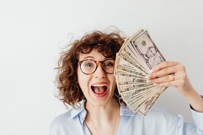 Woman with curly hair and glasses excitedly holding a fan of U.S. dollars, symbolizing random world facts and fresh perspectives.