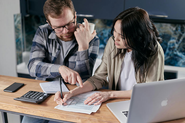 A couple discussing finances over a laptop, calculator, and documents at a kitchen table.