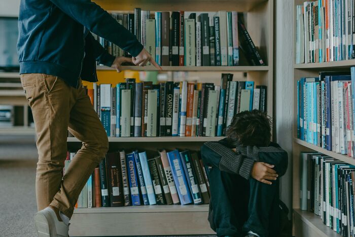 Person sitting by a library bookshelf, covering their face, experiencing a mic-drop-moment as someone points at them.