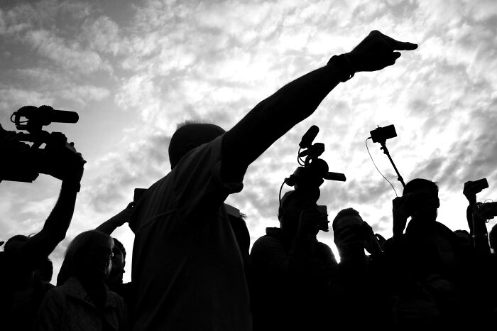 Silhouette of a person making a mic-drop gesture surrounded by cameras at sunset, capturing a dramatic moment.