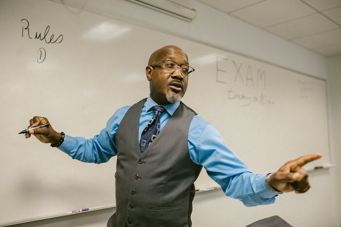 A teacher in a blue shirt and vest, marking a Mic-Drop-Moment with a gesture in front of a whiteboard in a classroom setting.