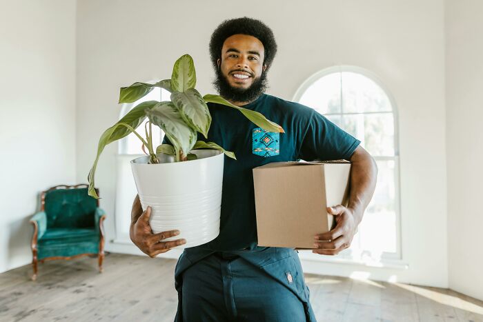 Man holding a potted plant and a box, smiling in a spacious room, illustrating aspects of childfree lifestyle.