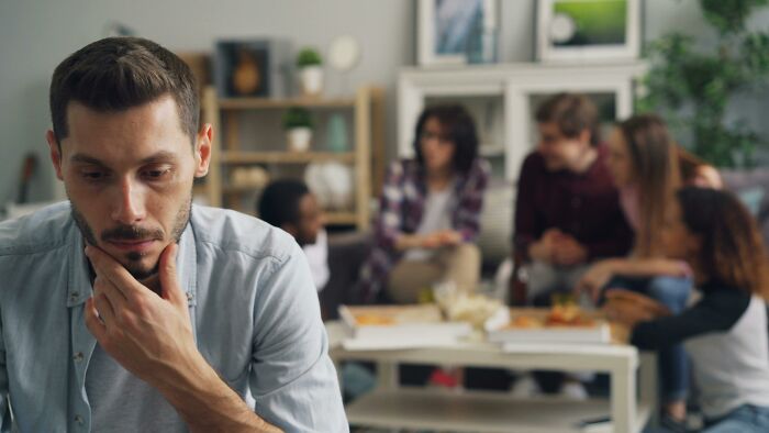 A thoughtful man sits alone while a group socializes in the background, highlighting childfree lifestyle choices.