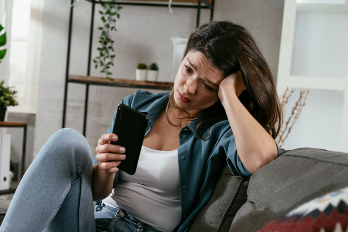 Woman sitting on couch, looking worried at a smartphone, contemplating a booked holiday vacation.