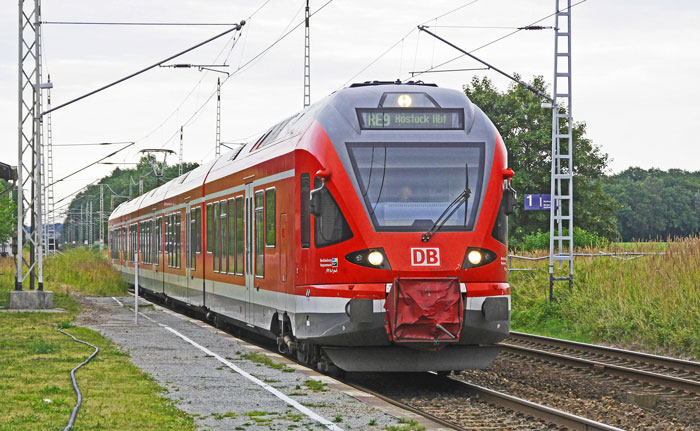 Red train on a railway track surrounded by greenery, under a cloudy sky.