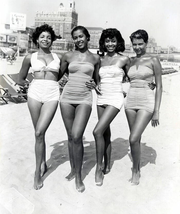 Four women in vintage swimsuits posing on a sunny beach, with historic buildings in the background.