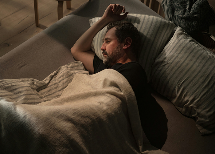 Man sleeping on a bed under a light blanket, arms folded, with gentle lighting in a cozy room.