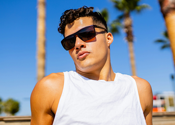 Man in sunglasses under a clear blue sky, wearing a white tank top, with palm trees in the background.