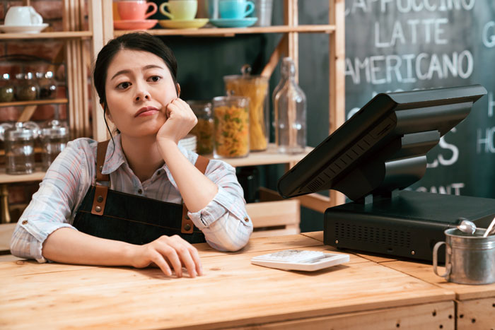 Restaurant worker covering shift, looking bored, sitting by a cash register, with coffee shop decor in the background.