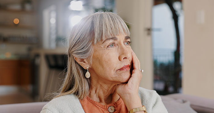 A contemplative older woman sitting on a sofa, wearing earrings and a watch, in a bright room.